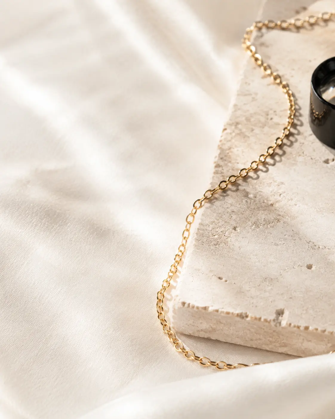 A delicate gold chain curves across ivory silk and pale limestone beside a jeweler's loupe.