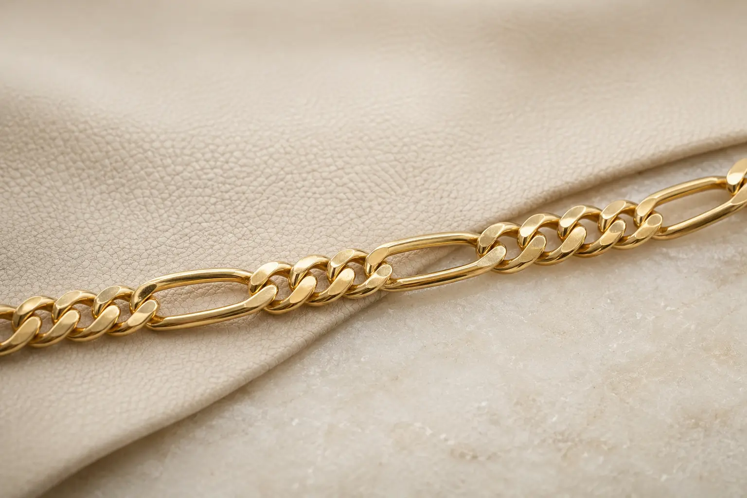 A gold Figaro chain with repeating short and elongated links crosses ivory leather and pale stone.