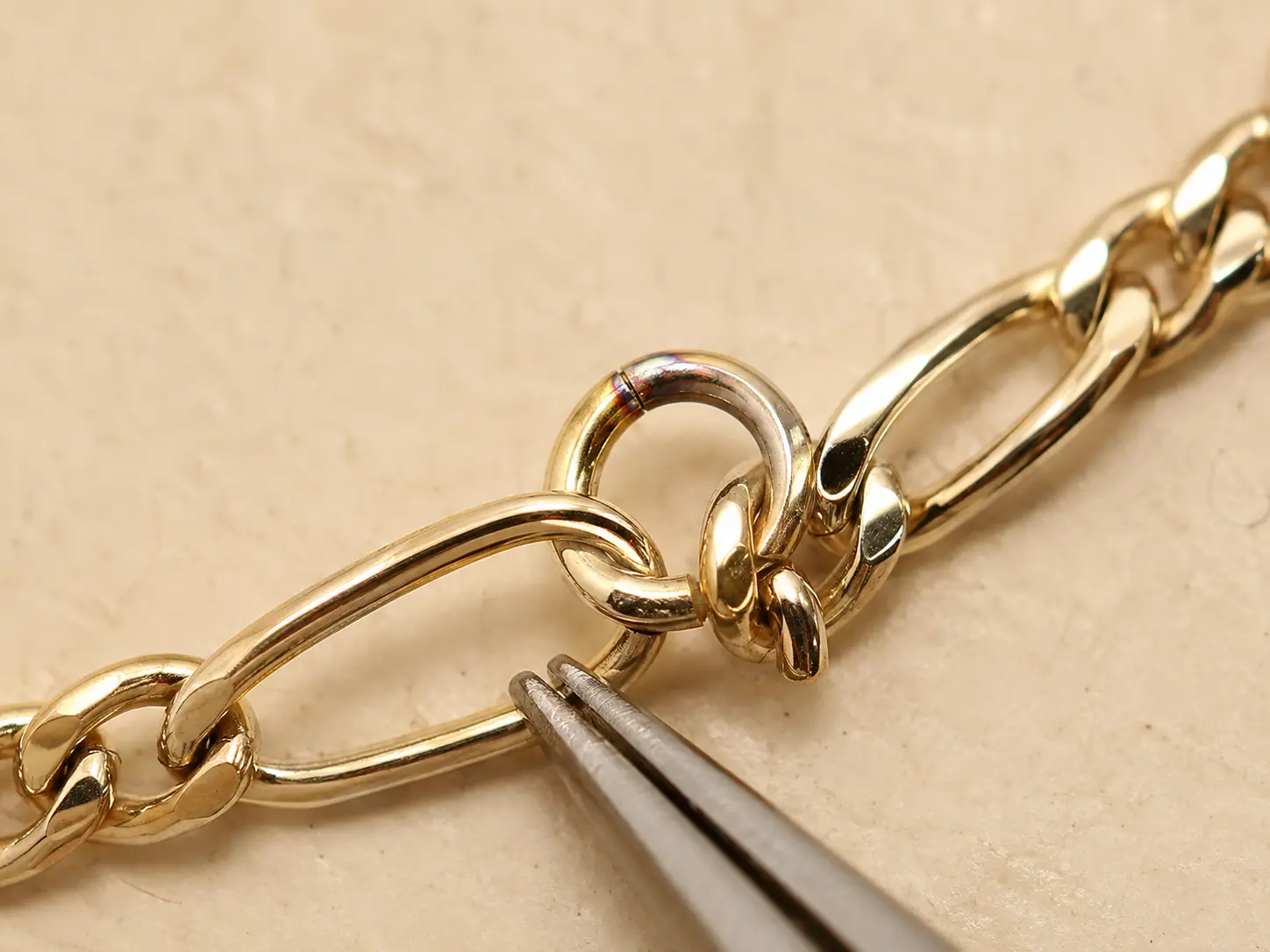An extreme macro shows a solder seam on a gold closure ring beside Figaro links and tweezers.